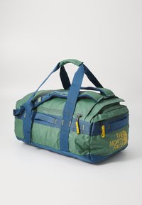 Duffle bag in green and blue fabric with yellow accents. Features top handles, a zippered pocket, and The North Face logo on the side.