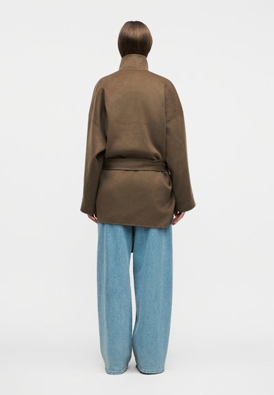 Persoană văzută de spate, purtând o jachetă maro cu curea și blugi largi, albastru deschis, pe un fundal alb simplu.