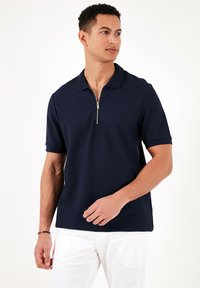Tricou polo bleumarin din țesătură texturată, cu un fermoar scurt la guler. Mâneci scurte și o etanșare dreaptă, asociat cu pantaloni albi.