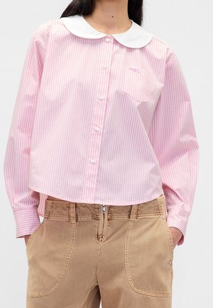 Person iført en pink og hvidstribet skjorte med knapper og en hvid rund krave samt beige bukser med forlommer og lynlås.
