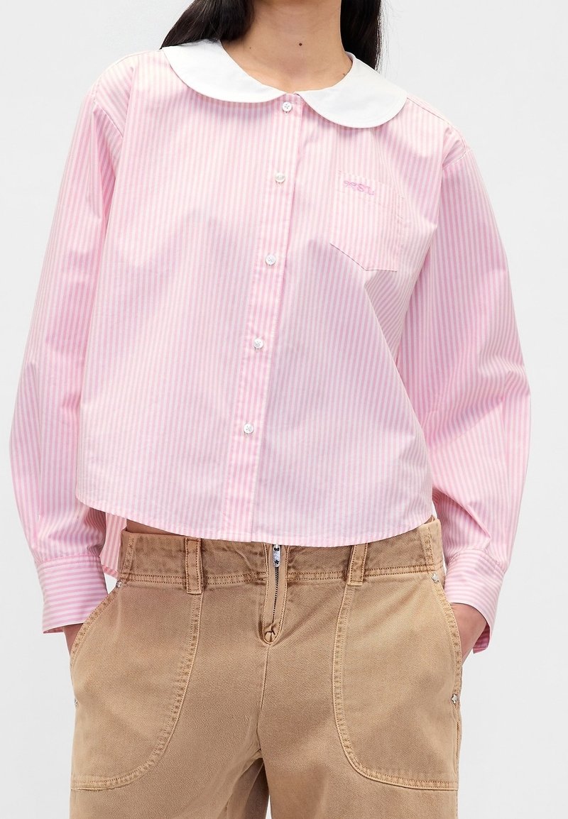 Personne portant une chemise rayée rose et blanche boutonnée avec un col rond blanc et un pantalon beige avec poches avant et fermeture éclair.