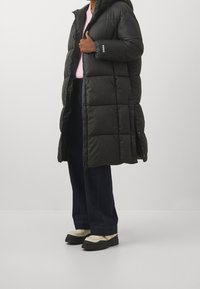 Lang sort pufferjakke med hætte, kviltet design, frontlynlås og trykknapper. Bæret over en pink sweater og parret med brede jeans.