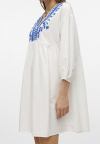 Vit bomullskjol med blå blommönsterbroderi, V-halsringning och trekvarts pösiga ärmar. A-line silhuett med rynkad midja.