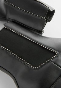 Gros plan sur des bottines en cuir noir avec des panneaux latéraux élastiques bordés de petits clous argentés.