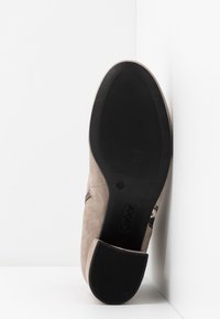 Chaussure en daim beige reposant sur la semelle contre un coin blanc, montrant une semelle noire texturée avec le nom de la marque "Gabor" et les détails du talon.