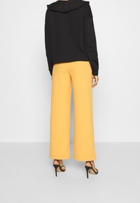 Felpa nera con un colletto arricciato, abbinata a pantaloni gialli a gamba larga. L'outfit è completato da sandali neri con tacco alto.