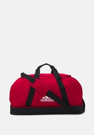 Sac de sport en tissu rouge avec des poignées et une sangle noires, arborant un logo Adidas blanc sur le devant. Forme arrondie avec une fermeture éclair.