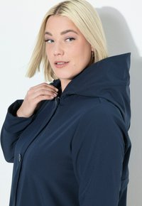 Giacca con cappuccio blu navy realizzata in tessuto testurizzato, dotata di chiusura frontale con bottoni e colletto alto, che offre una silhouette elegante.