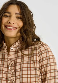 Blouse en flanelle beige avec col à volants, boutons clairs et texture douce. Présente des motifs à carreaux marron et beige, une coupe ajustée et des manches longues.