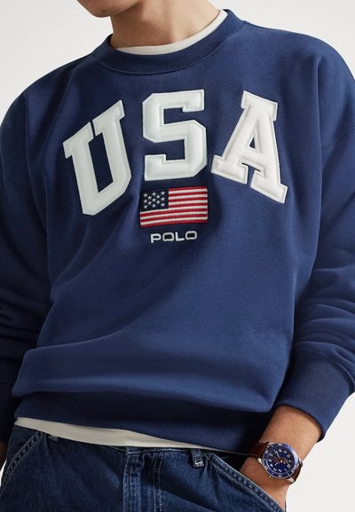 Μπλε σκούρο φούτερ με κέντημα "USA" και μία μικρή αμερικανική σημαία στο στήθος, στρογγυλή λαιμόκοψη και απαλό βαμβακερό ύφασμα. Φοριέται με τζιν.