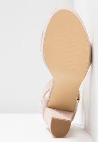 Béžové semišové sandály s blokovým podpatkem, ukazující hladkou béžovou podešev a texturovaný špičkový podpatek na bílém pozadí.