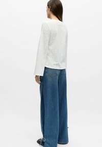 Kvinne iført hvit langermet topp med blomstertekstur og vide blå jeans med snøring på sidene, stående mot hvit bakgrunn.