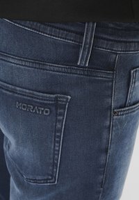 Tamne traperice s ušivenim detaljima, koje imaju stražnji džep s utisnutim natpisom "MORATO" i malim metalnim akcentom pored džepa.