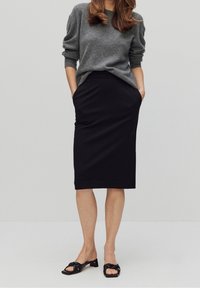 Pull gris en tricot avec des manches amples, associé à une jupe crayon noire arrivant au genou. Des sandales à talons blocs noires complètent la tenue.