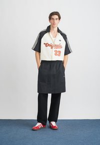 Svart og hvit skjorte med krage med rød "Originals 33" trykk, kombinert med svarte bukser og røde joggesko. Glatt stoff, uformelt design.