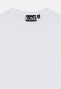 Bela majica s crew izrezom in majhnim zlatim logotipom "EA7 Emporio Armani" na prsih ter črnimi oznakami velikosti pri ovratu.