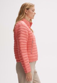 Doudoune rose avec un col haut, design matelassé, poche zippée sur le côté et texture lisse, portée sur un pantalon beige.