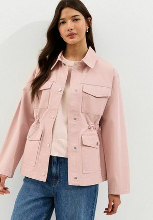 Donna che indossa una giacca rosa chiaro con bottoni e quattro tasche frontali sopra un maglione beige e jeans blu, in piedi contro un muro bianco.