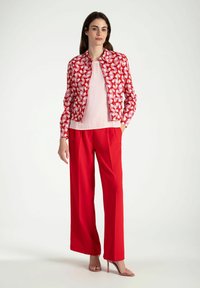 Rote Jacke mit einem Muster aus pinken und weißen geometrischen Designs, kombiniert mit einem hellrosa Top und weit geschnittenen roten Hosen. Das Model trägt High Heels.