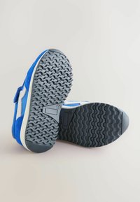 Niebieskie buty sportowe z teksturowaną, szarą gumową podeszwą, z białymi akcentami i rzepem. Cholewka wykonana jest z połączenia zamszu i tkaniny.