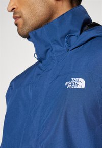 Mėlyna striukė iš tekstūruoto audinio, su aukštu apykakle ir balta "The North Face" logotipu ant krūtinės.