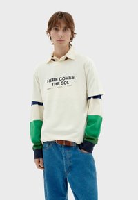 Camisa de manga larga de color crema claro con el texto "AQUÍ VIENE EL SOL", con mangas en bloques de color verde, azul y crema. Llevada con jeans azules.
