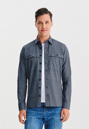 Szara koszula na guziki z kołnierzykiem, dwoma kieszeniami na piersiach i luźnym fasonem, noszona na białej koszulce i zestawiona z niebieskimi dżinsami.