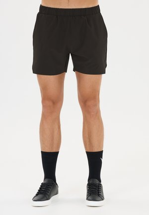 Mannelijke benen met zwarte sportshorts, zwarte sokken tot halverwege de kuit en zwarte sneakers met witte zolen op een witte achtergrond.