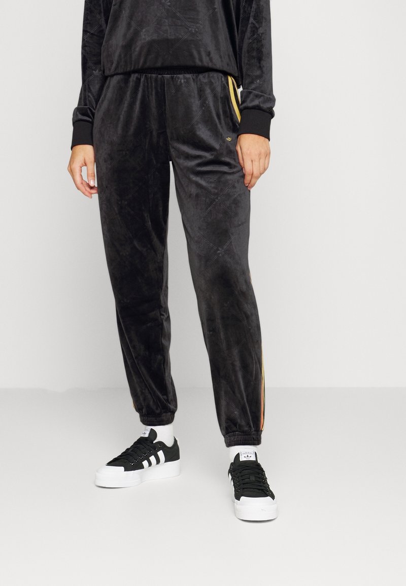 Schwarze, samtige Jogginghosen mit lockerer Passform, ausgestattet mit elastischen Bündchen und seitlichen Streifen in Gold. Kombiniert mit schwarzen Sneakers.