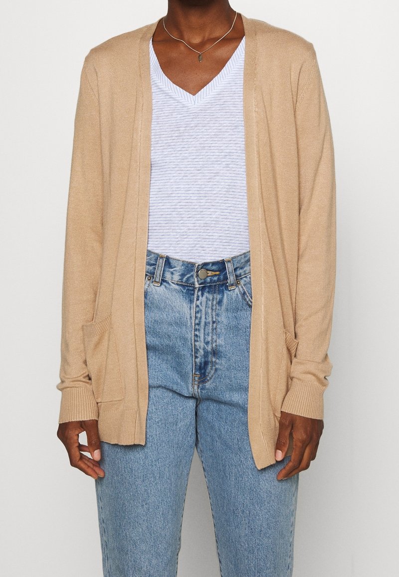 Personne portant un t-shirt à col en V rayé bleu clair, un cardigan beige ouvert à l'avant avec des poches, un jean en denim bleu taille haute, et un collier argenté.