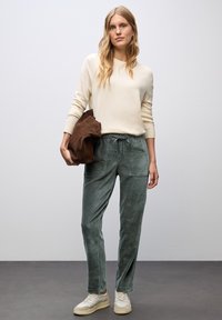 Kvinna med långt blont hår som bär en krämfärgad tröja, gröna byxor med dragsnöre, beige sneakers, och håller en brun mockajacka under armen.