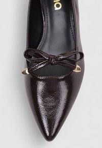 Zapato de tacón alto de punta afilada en piel sintética brillante de color marrón oscuro. Presenta un lazo decorativo y acentos de hardware en tono dorado. Textura suave.