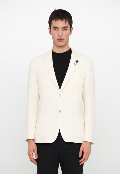 Blazer beige avec deux poches avant, trois boutons et une épingle florale noire. Porté sur un col roulé noir à rayures. Texture lisse.