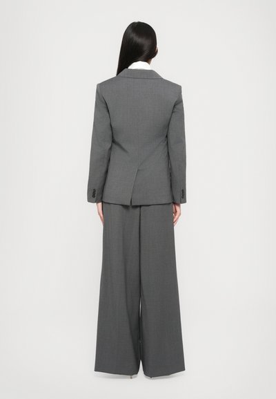 Donna con i capelli lunghi e lisci indossa un blazer grigio su misura e pantaloni larghi, in piedi di fronte a uno sfondo bianco semplice.