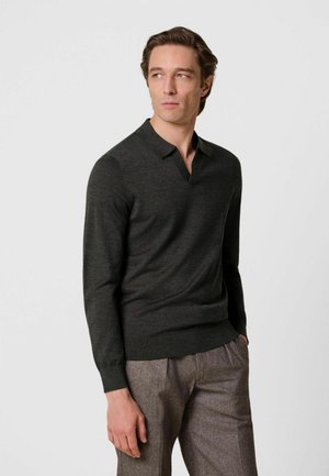 Man wearing dark long-sleeve polo shirt and brown trousers, standing with one hand in pocket, looking to his left against a plain background.