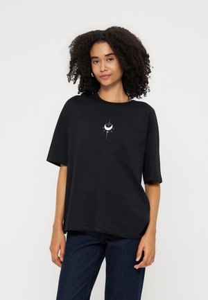 Femme aux cheveux bouclés portant un t-shirt noir ample avec un petit motif blanc de croissant de lune et d'étoiles, associé à un jean bleu foncé.