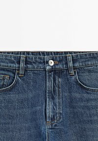 Vue de face de la taille d'un jean en denim bleu avec fermeture par bouton et passants de ceinture sur fond blanc.