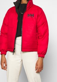 Rød pufferjakke med lynlås fortil, elastiske manchetter og en sort "Helly Hansen" logo. Materialet er skinnende og isoleret for varme.