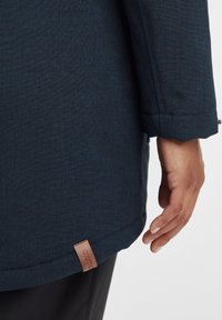 Granatowy płaszcz z teksturowanej tkaniny, z skórzaną metką na dolnym końcu i mankietem na prawej stronie.