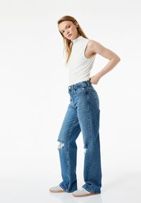 Top blanco sin mangas ligero combinado con jeans rotos de tiro alto en azul, con un ajuste relajado y detalles desgastados.