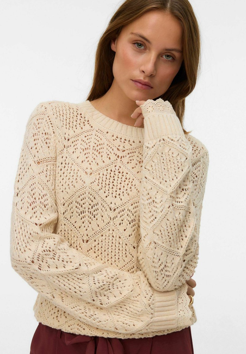 Femme aux cheveux châtain clair portant un pull en tricot crème avec des motifs en diamant et des poignets en dentelle, posant avec la main près du menton sur un fond blanc.