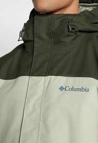 Veste imperméable vert olive et vert clair avec un col haut, dotée de matériel ajustable et du logo Columbia en bleu sur le devant.