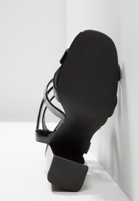 Sandale à talons hauts noire avec finition brillante, design ouvert et talon épais; avec fines brides autour de la cheville et semelle texturée.