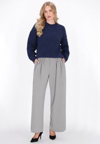 Granatowy sweter prążkowany z długim rękawem, w połączeniu z jasnoszarymi, plisowanymi spodniami w szerokim kroju oraz czarnymi szpilkami. Modelka stoi pewnie.