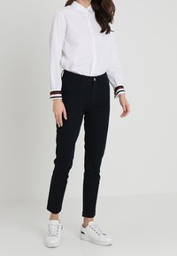 Vit skjorta med knäppning framtill och randiga manschetter, kombinerad med mörka slim fit-byxor och vita sneakers med accentdetaljer.