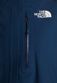 Veste bleu foncé avec une poche poitrine verticale zippée et le logo blanc The North Face au-dessus de la poche du côté droit.