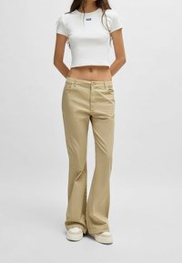 Vit kortärmad t-shirt med en liten blå logotyp, kombinerad med beige utsvängda byxor och vita sneakers. Enkel textur och avslappnad passform.