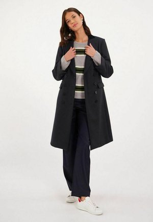 Manteau noir croisé en mélange de laine, avec un col cranté, associé à un pull à rayures et un pantalon bleu marine, complété par des baskets blanches.