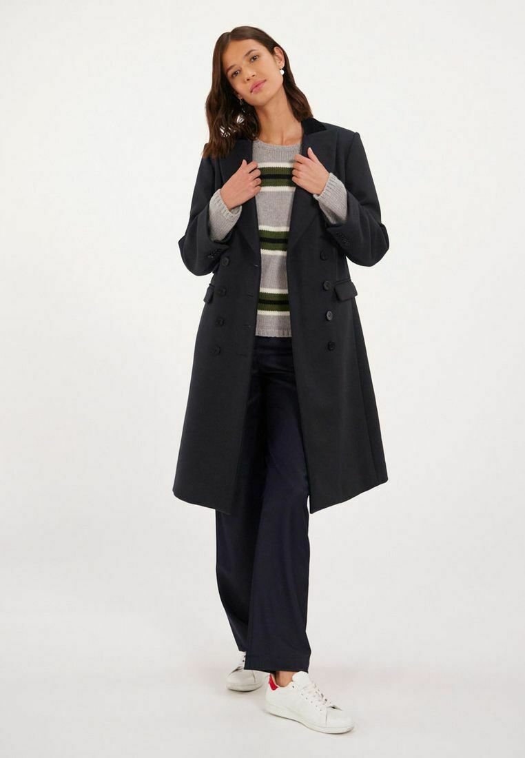 Manteau noir croisé en mélange de laine, avec un col cranté, associé à un pull à rayures et un pantalon bleu marine, complété par des baskets blanches.