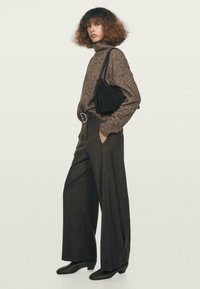 Hnědý pletený rolák, tmavé kalhoty s vysokým pasem a širokými nohavicemi, a černá taška. Modelka nosí černé špičaté kotníkové boty. Minimalistické pozadí.